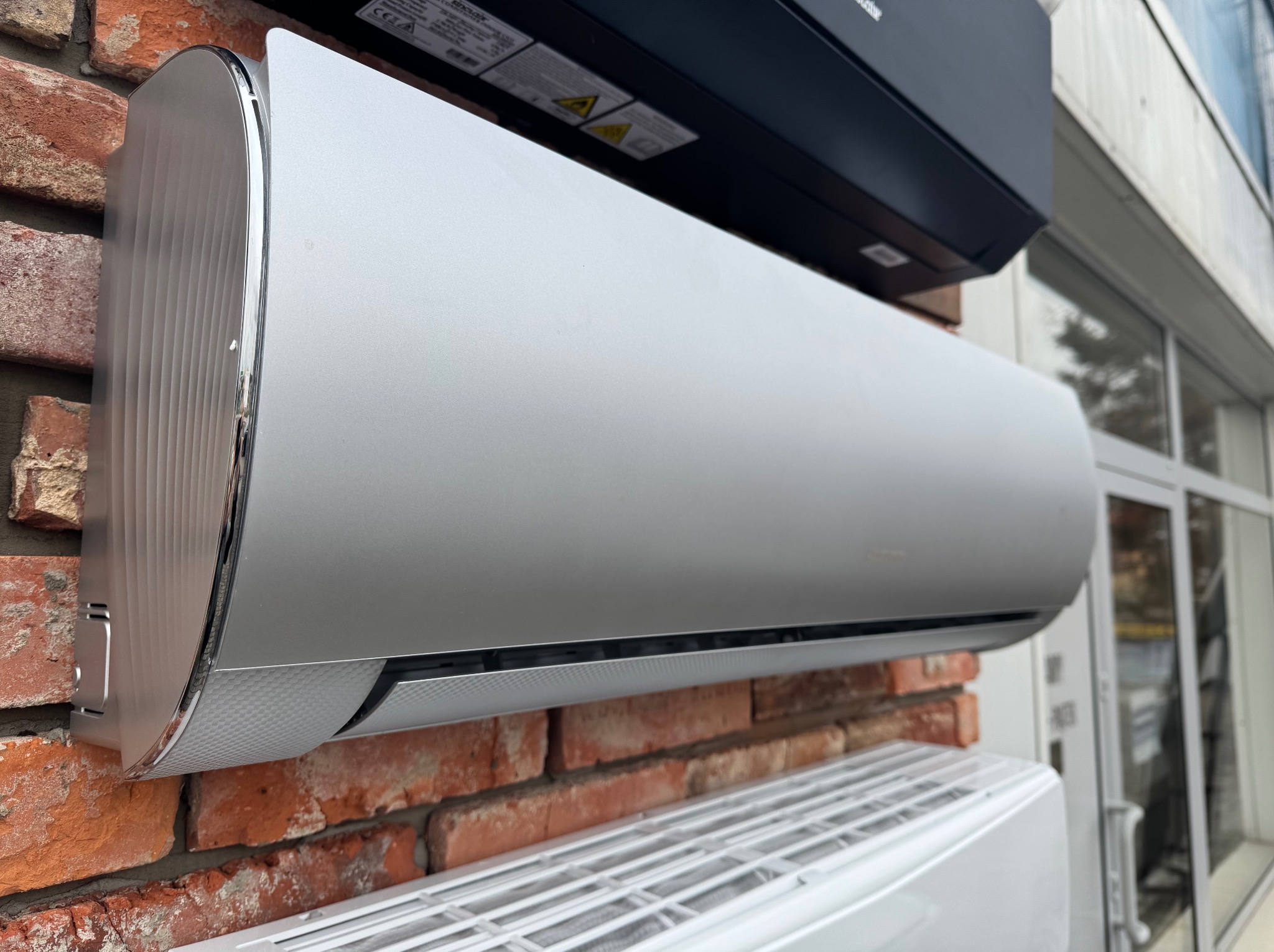 Srebrny klimatyzator ścienny na tle ceglanej ściany, widoczne inne jednostki klimatyzacyjne powyżej i poniżej, obok przeszklone drzwi.
