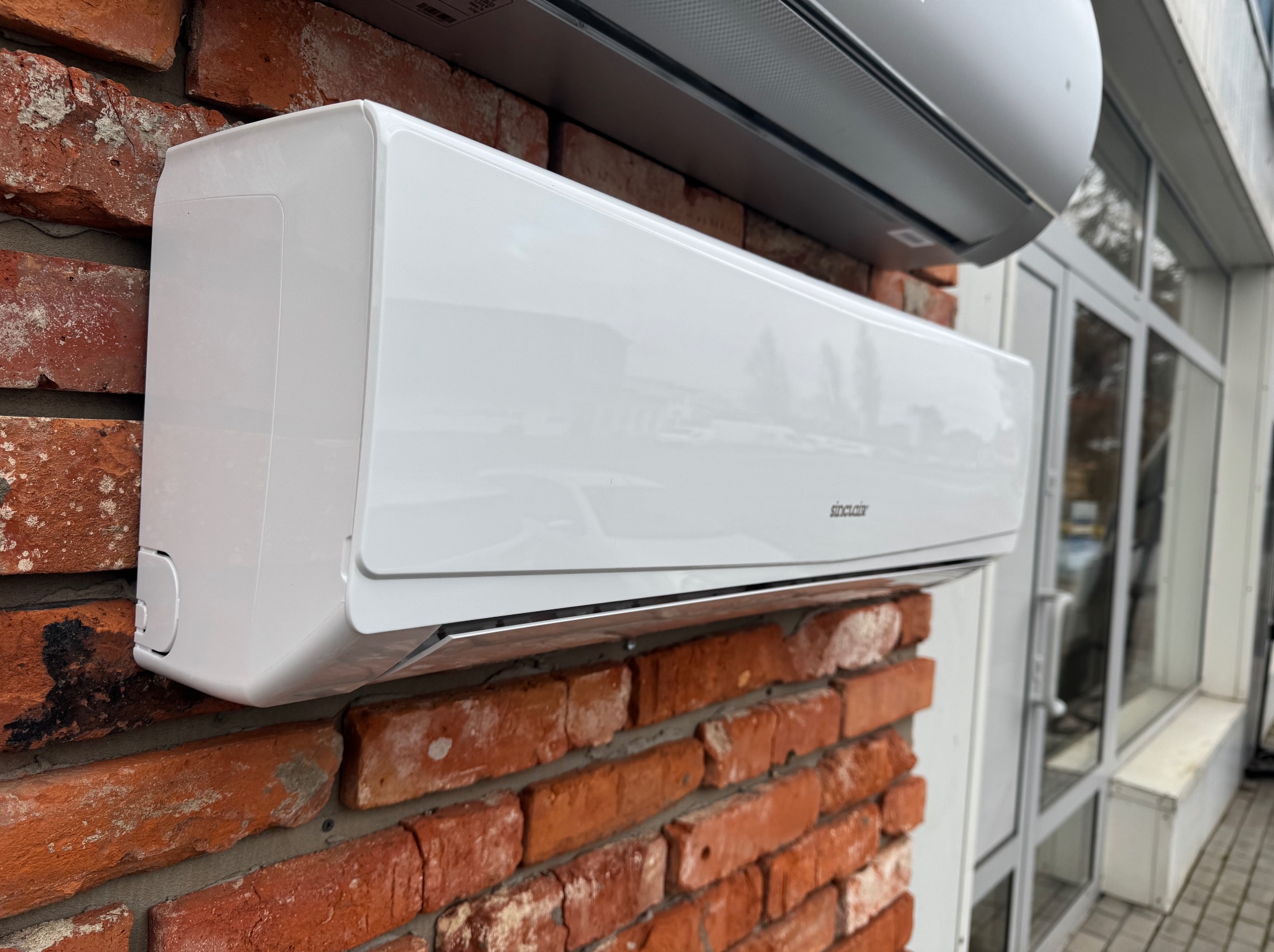Biała jednostka wewnętrzna klimatyzacji Sinclair zamontowana na ceglanej ścianie, widoczna z boku, z drugą jednostką na górze i oknami w tle.