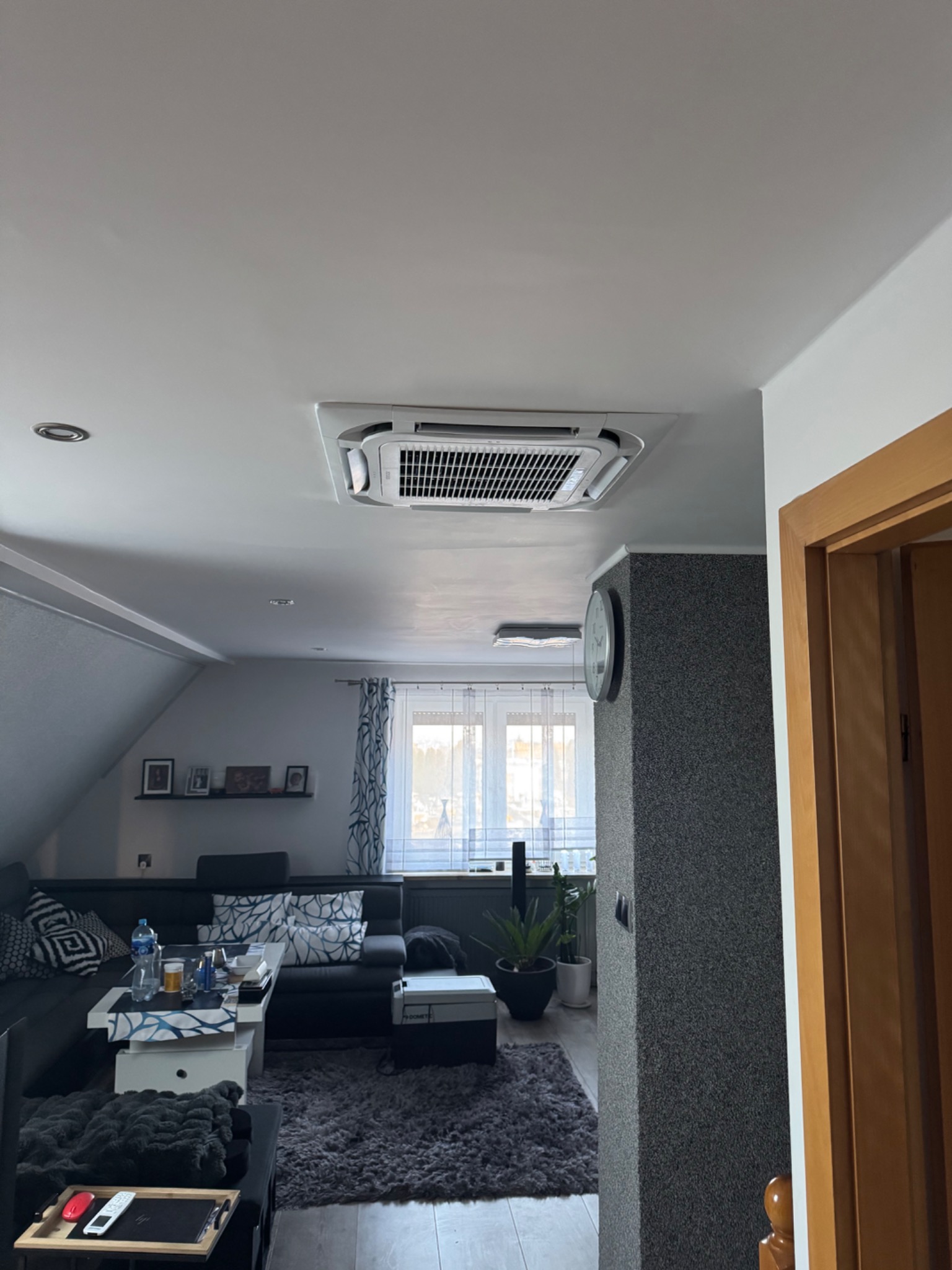 Kasetonowa jednostka klimatyzacji zamontowana w suficie poddasza mieszkalnego, widok na część salonu z szarą sofą i oknem.