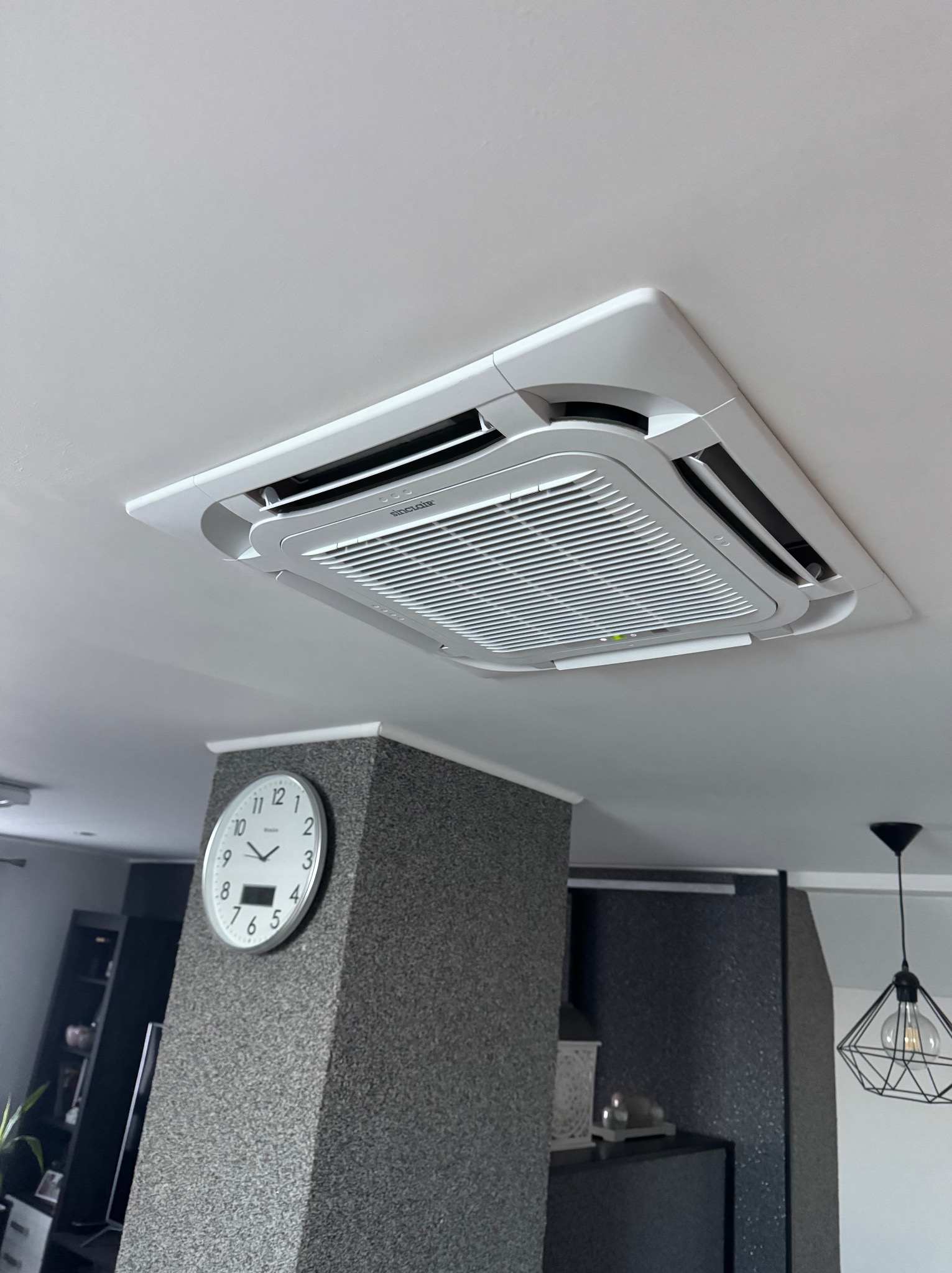 Kasetonowa jednostka klimatyzacji sufitowej marki Sinclair w nowoczesnym wnętrzu z widocznym zegarem ściennym i lampą wiszącą.