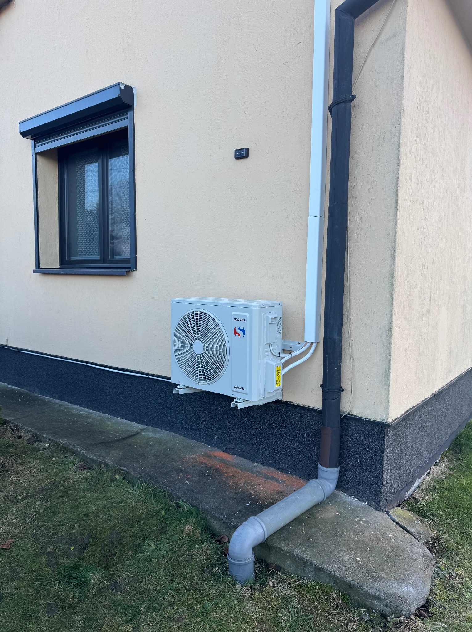 Biała jednostka zewnętrzna klimatyzacji zamontowana na elewacji domu w pobliżu okna z roletą i rynny spustowej.