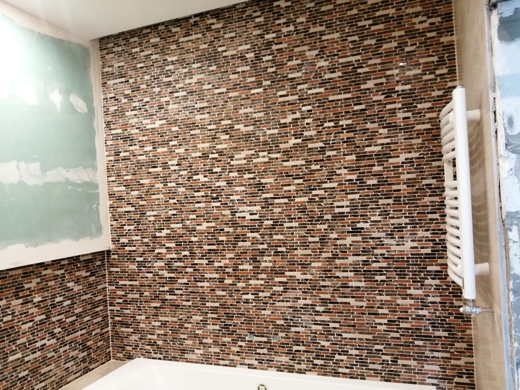 Mozaika z małych, prostokątnych płytek w odcieniach brązu, beżu i czerni na ścianie w łazience, z widocznym białym grzejnikiem drabinkowym i fragmentem ściany przygotowanej pod malowanie.