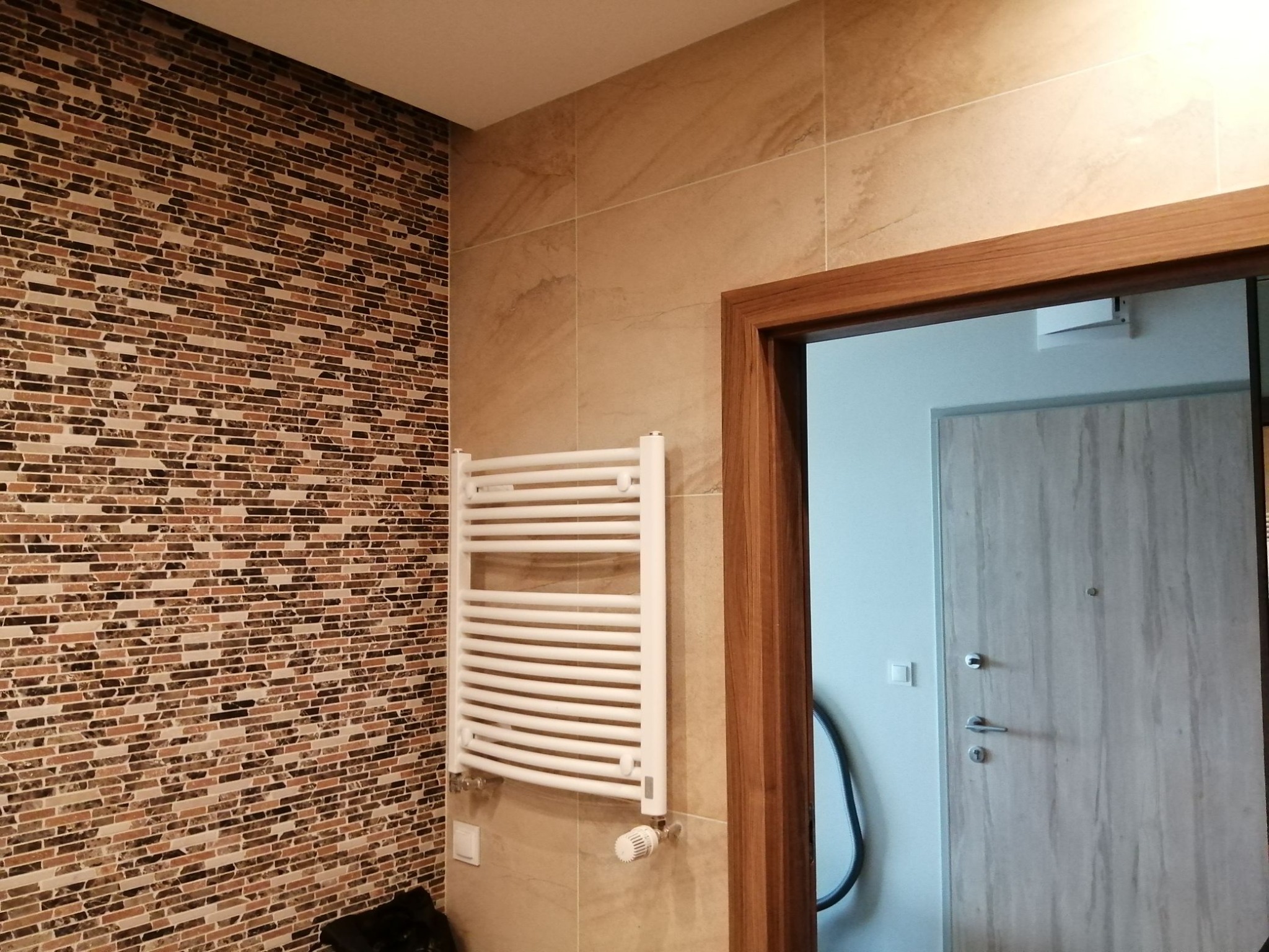 Wykończona łazienka w Warszawie z mozaikową ścianą, białym grzejnikiem drabinkowym i fragmentem drewnianej futryny drzwiowej.