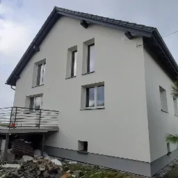 Ocieplony i pomalowany na biało dom jednorodzinny z szarą podmurówką i ciemnoszarą dachówką, widoczny z boku, z balkonem i stertą gruzu u podstawy.