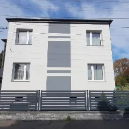 Dwupiętrowy dom z nową, biało-szarą elewacją, widoczny zza szarego, poziomego ogrodzenia, z zaparkowanym obok samochodem dostawczym.