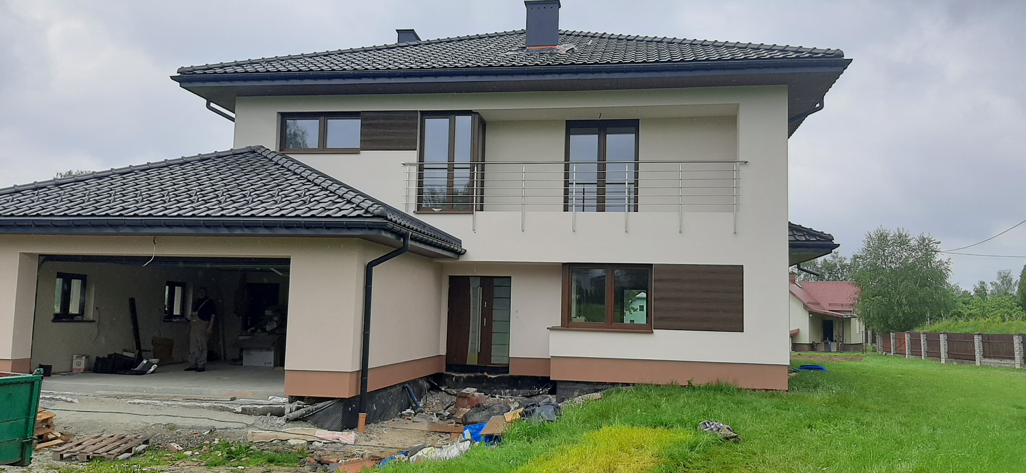 Dwukondygnacyjny dom jednorodzinny z garażem w trakcie wykończenia elewacji, widoczne rusztowania i materiały budowlane wokół budynku.