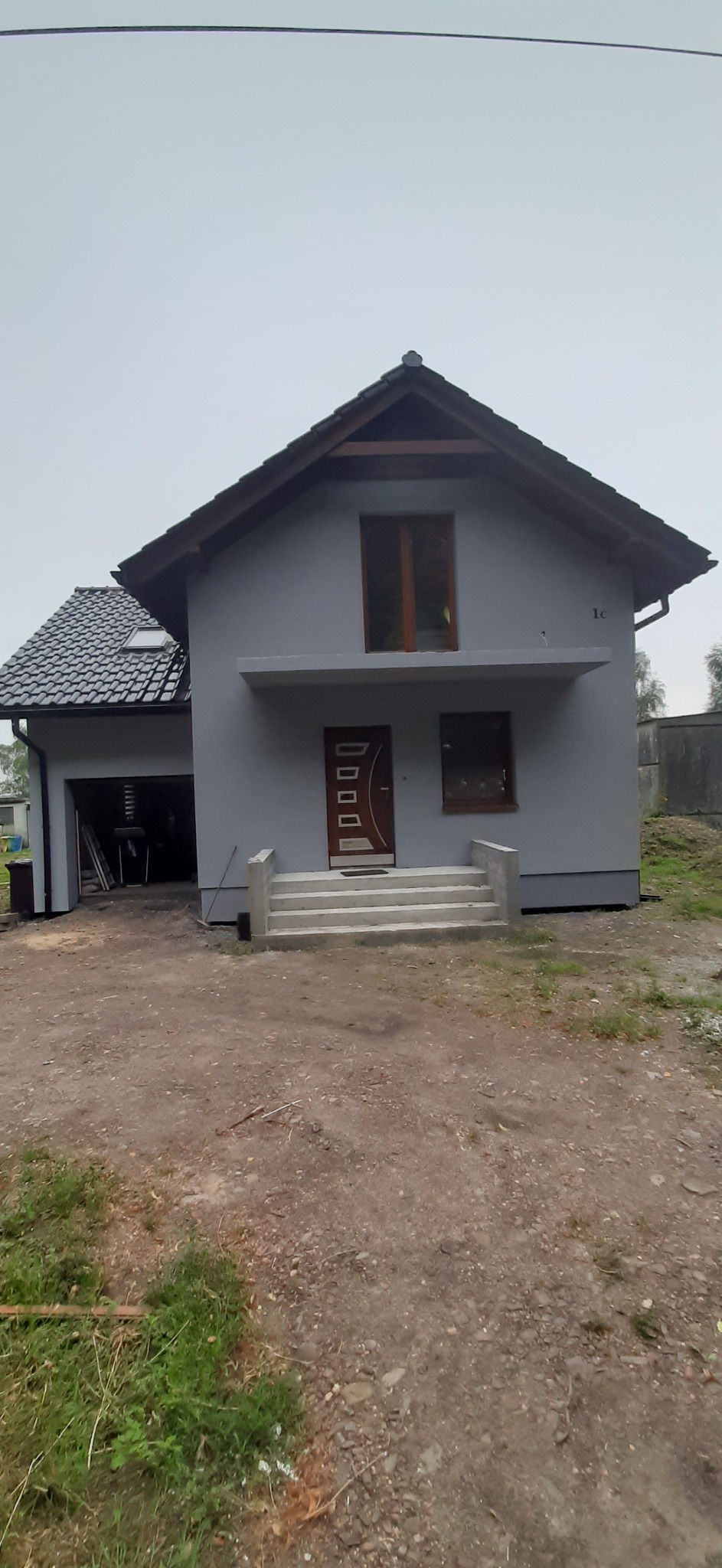 Szary dom jednorodzinny z ciemną dachówką, brązowymi oknami i drzwiami wejściowymi z elementami dekoracyjnymi, widok z zewnątrz na niezagospodarowaną działkę.