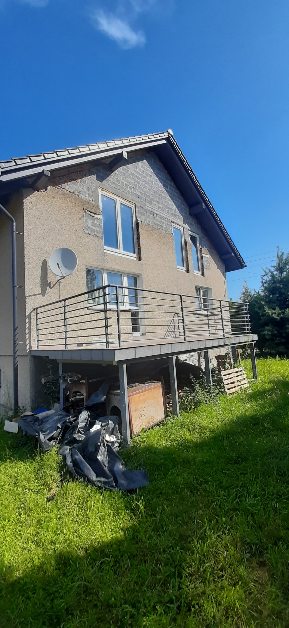 Dom z szarą elewacją w trakcie prac, z widocznymi fragmentami ocieplenia i niezabezpieczonymi oknami, balkon z metalową balustradą, antena satelitarna na ścianie, nieporządek pod balkonem.