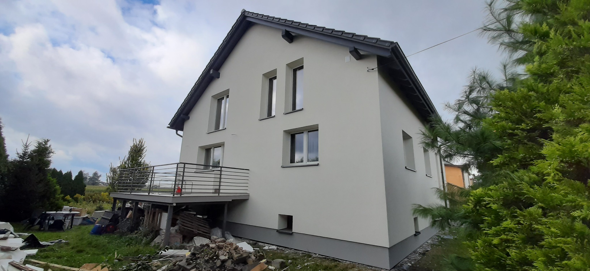 Ocieplony i pomalowany na biało dom jednorodzinny z szarą podmurówką i ciemnoszarą dachówką, widoczny z boku, z balkonem i stertą gruzu u podstawy.