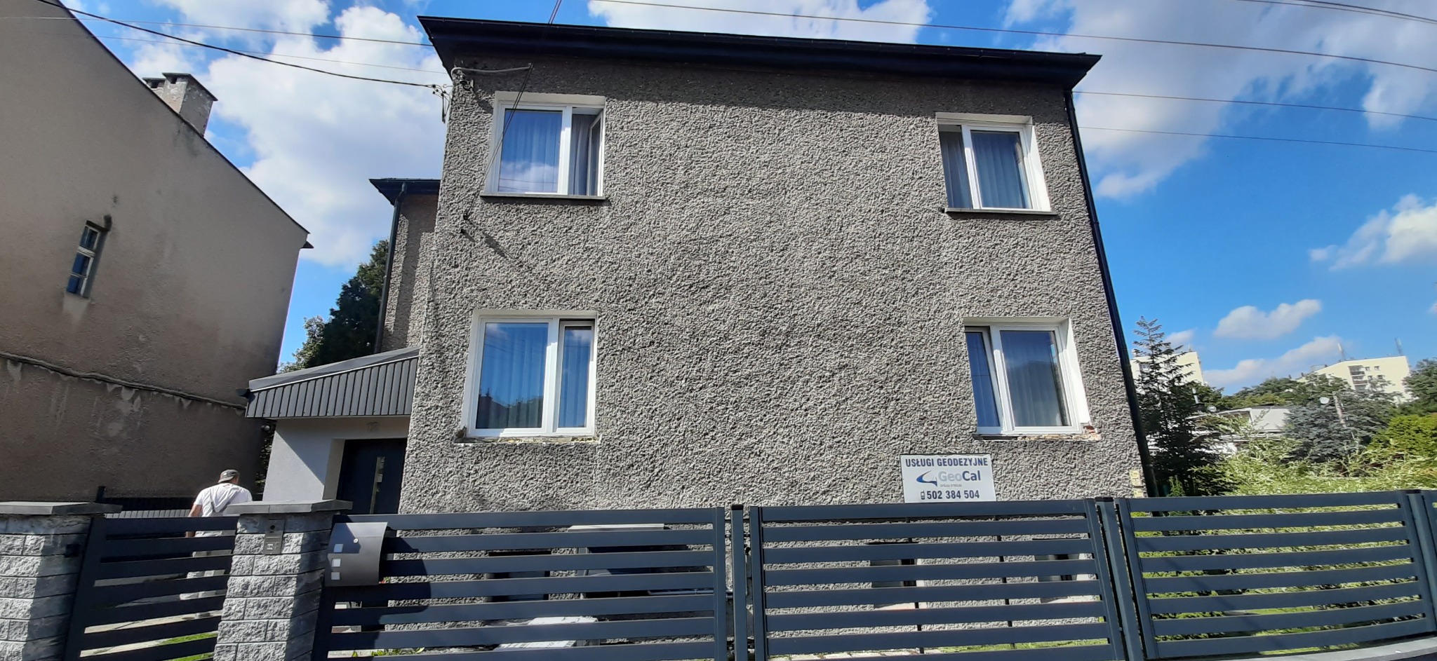 Szary dom dwupiętrowy z oknami w białych ramach, widoczny zza grafitowego ogrodzenia panelowego, z tabliczką informacyjną o usługach geodezyjnych.