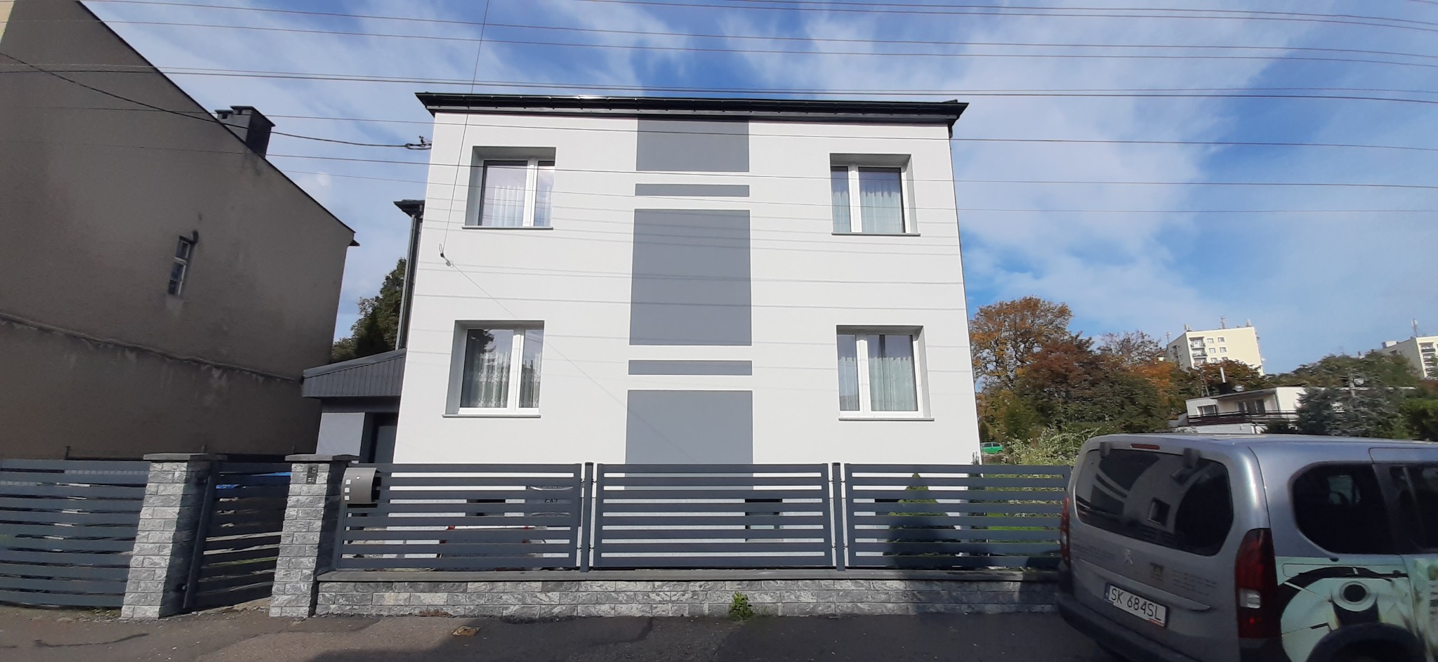 Dwupiętrowy dom z nową, biało-szarą elewacją, widoczny zza szarego, poziomego ogrodzenia, z zaparkowanym obok samochodem dostawczym.