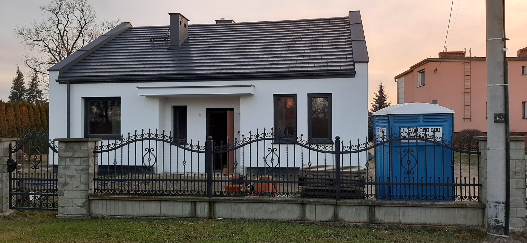 Nowy dom jednorodzinny z białą elewacją i ciemną dachówką, ogrodzony ozdobnym, kutym ogrodzeniem, z widoczną toaletą przenośną z boku.