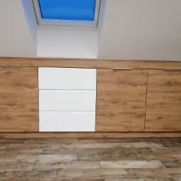 Niska komoda z frontami imitującymi drewno i białymi szufladami, umieszczona pod oknem dachowym, na podłodze imitującej drewniane deski.