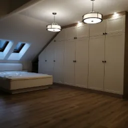 Sypialnia na poddaszu z zabudową szaf wnękowych pod skosem, drewniana podłoga i dwa okna dachowe.