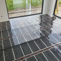 Montaż folii grzewczej pod podłogę w nowym domu, widoczne okno z widokiem na ogród, folia marki Termofol ułożona na całej powierzchni.