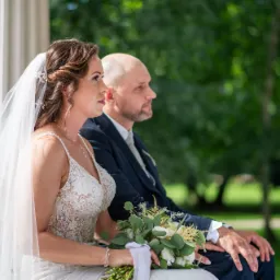 Panna młoda w koronkowej sukni i welonie, trzymająca bukiet, siedzi obok pana młodego w granatowym garniturze podczas ceremonii ślubnej na tle zieleni.