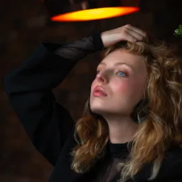 Portret kobiety z blond lokami w czarnej marynarce, spoglądającej w górę, z ręką na włosach, pod ciepłym światłem lampy, z rośliną doniczkową w tle.