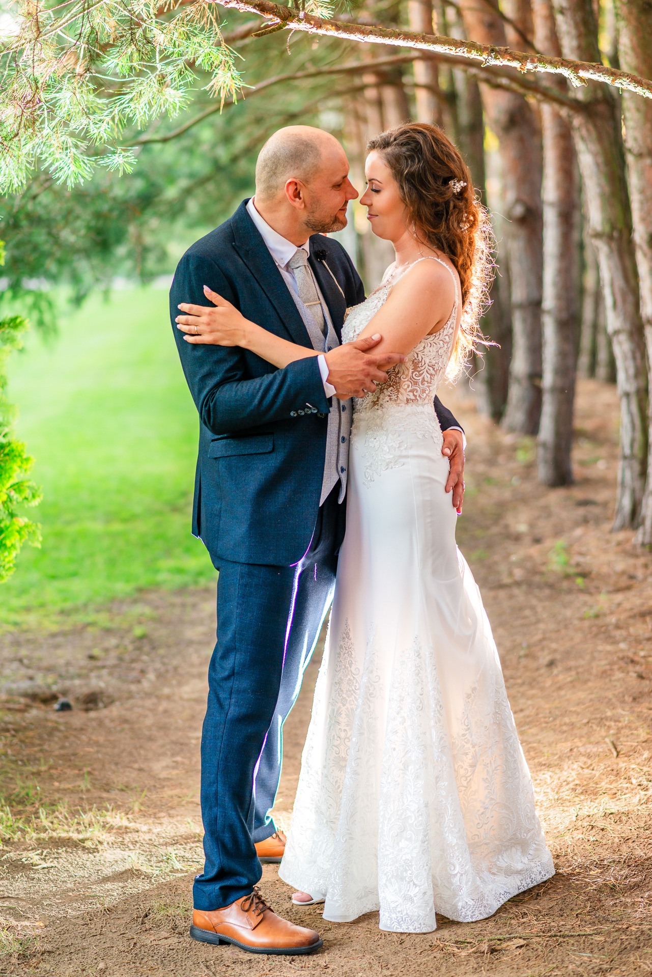 Para młoda w objęciach na leśnej ścieżce, panna młoda w koronkowej sukni z odsłoniętymi ramionami, pan młody w granatowym garniturze i szarej kamizelce, drzewa w tle.