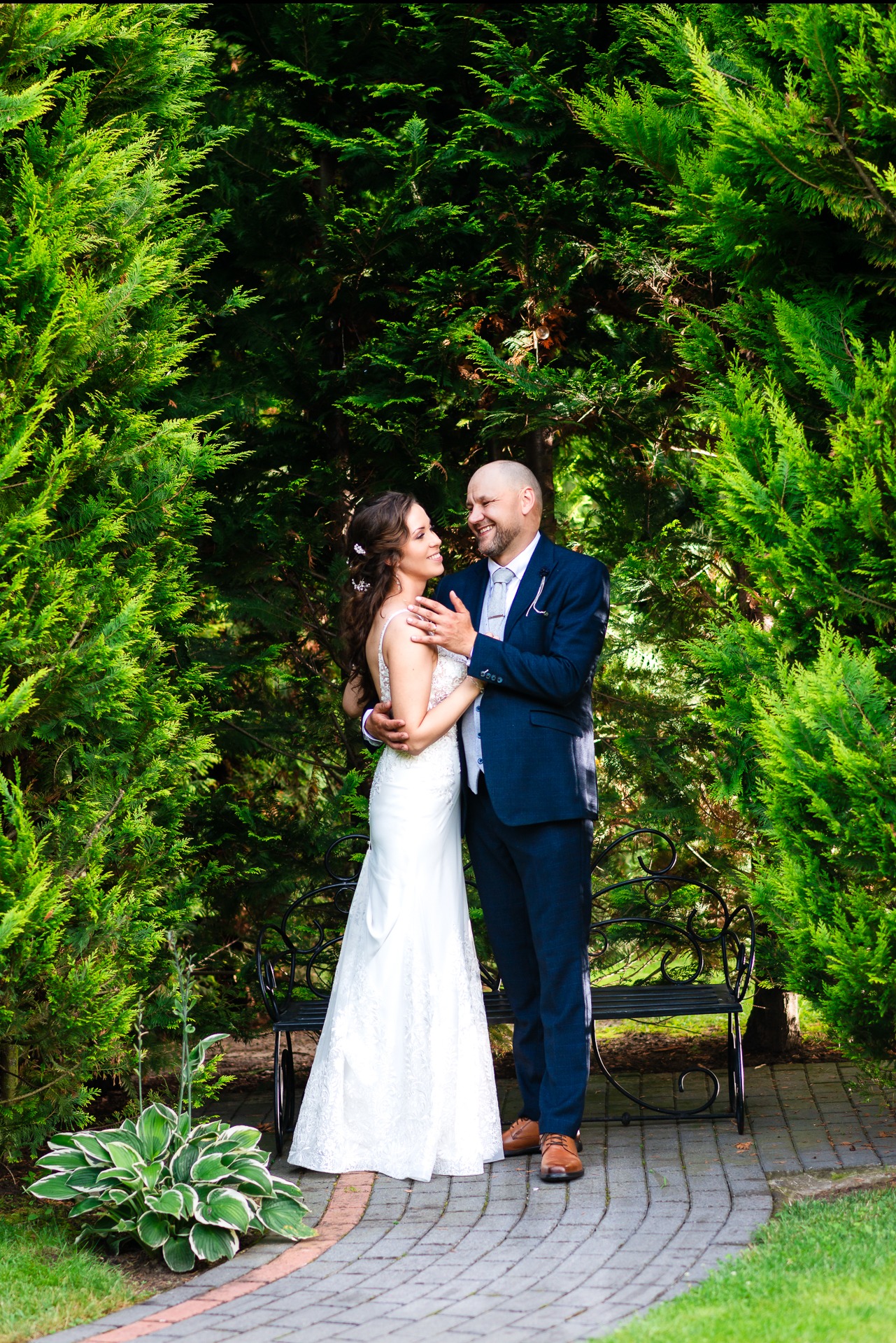 Para młoda, ona w białej koronkowej sukni z odkrytymi ramionami, on w granatowym garniturze i jasnym krawacie, obejmują się na tle zielonych krzewów i ozdobnej ławki w ogrodzie, oboje uśmiechnięci.