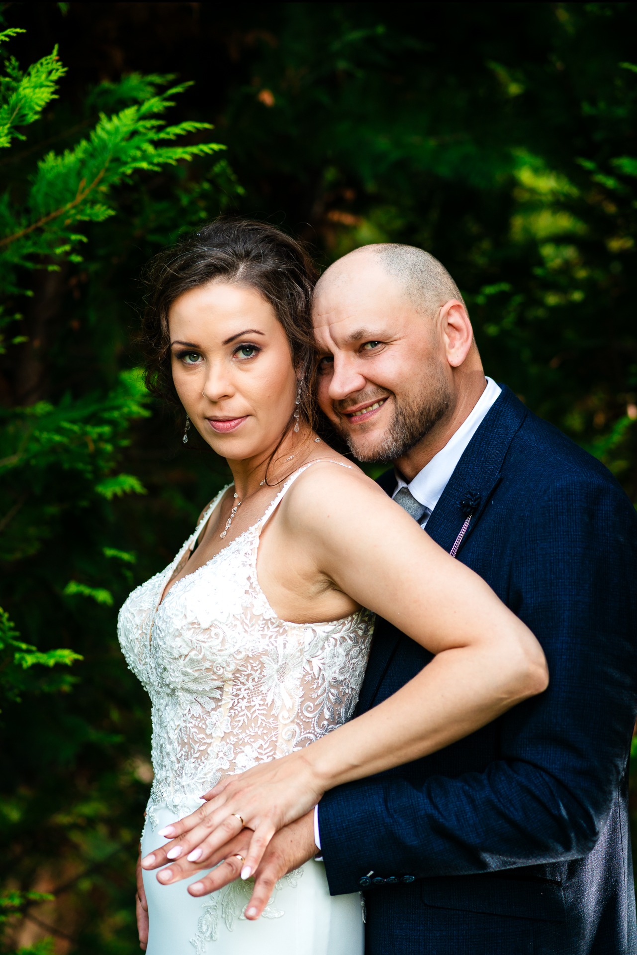 Uśmiechnięta para młoda w objęciach na tle zieleni: panna młoda w koronkowej sukni z odkrytymi ramionami i delikatną biżuterią, pan młody w granatowym garniturze z szarym krawatem.