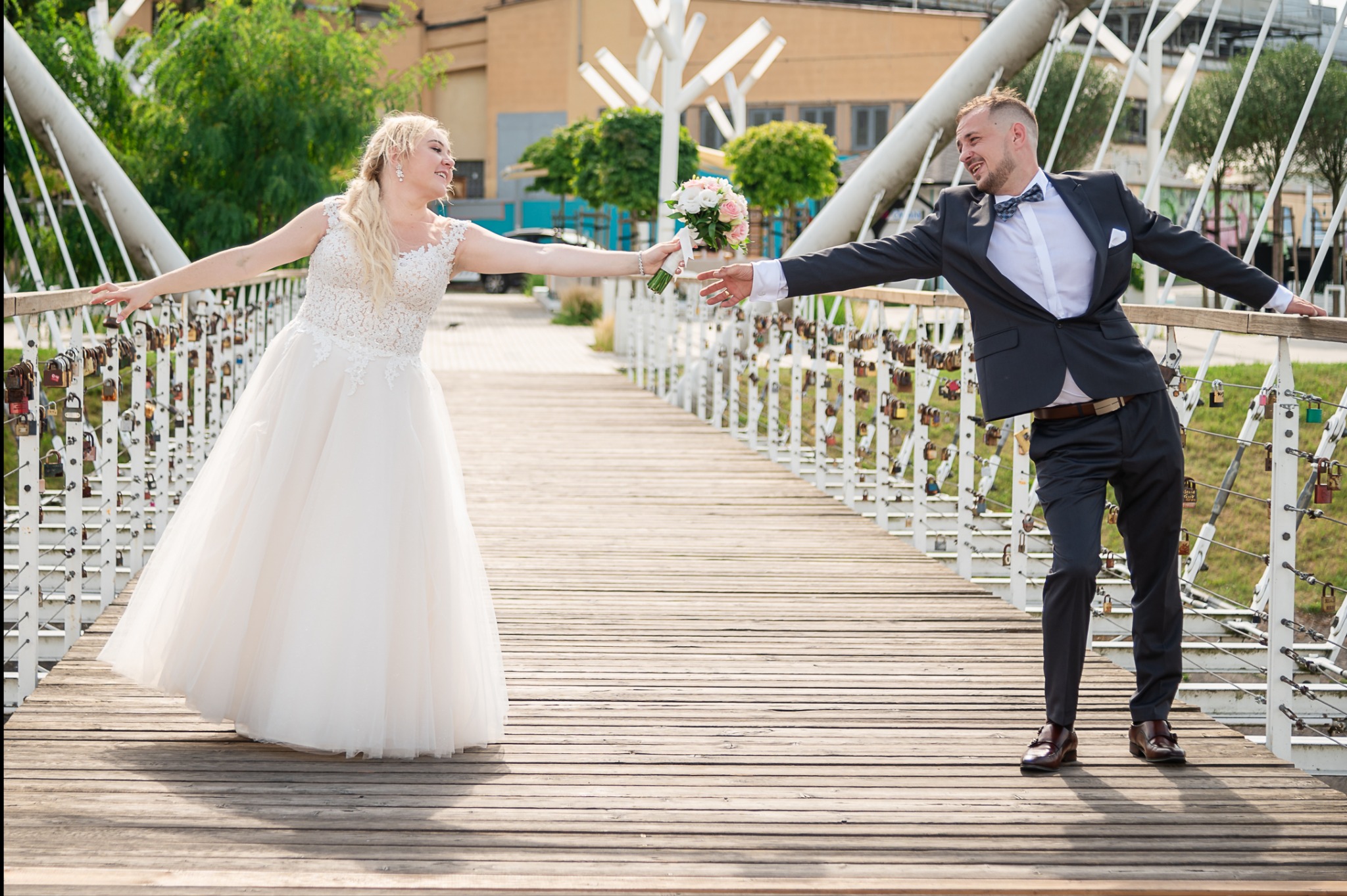 Para młoda na moście, panna młoda w koronkowej sukni ślubnej z bukietem, pan młody w garniturze z muszką, oboje opierają się o barierki mostu ozdobione kłódkami.
