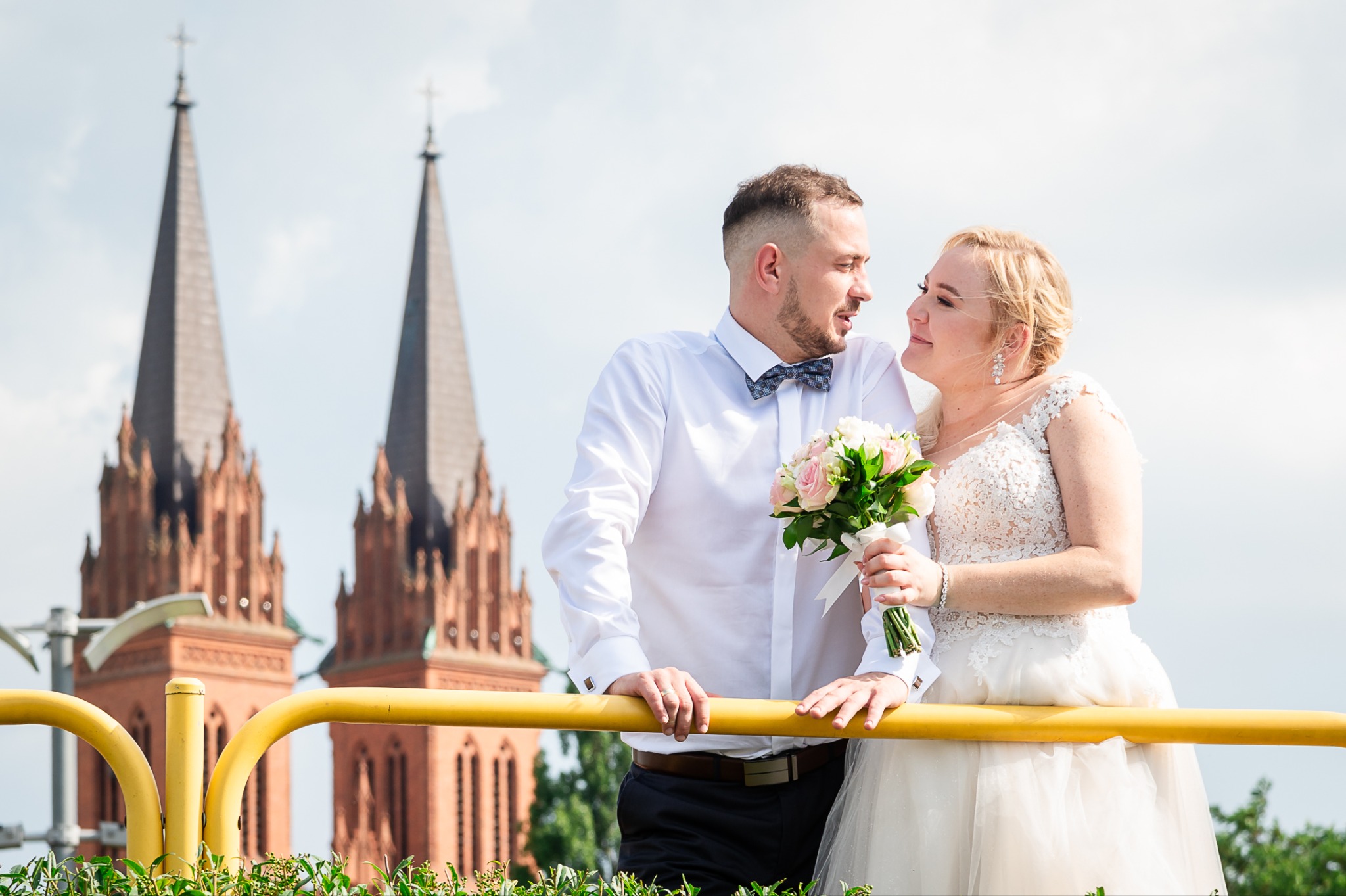Para młoda wpatrzona w siebie, opierająca się o żółtą barierkę, w tle wieże kościoła z czerwonej cegły i błękitne niebo.