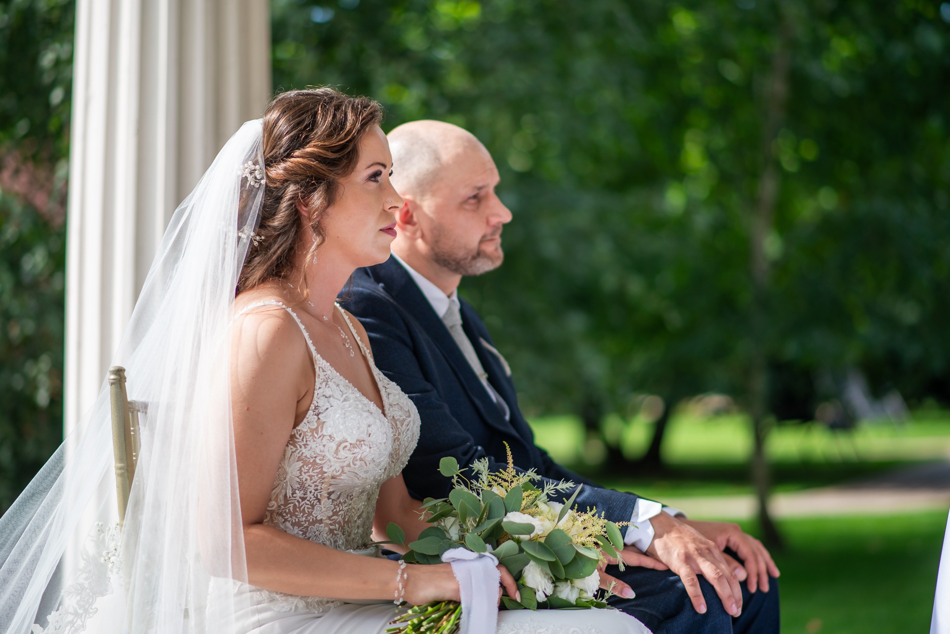 Panna młoda w koronkowej sukni i welonie, trzymająca bukiet, siedzi obok pana młodego w granatowym garniturze podczas ceremonii ślubnej na tle zieleni.