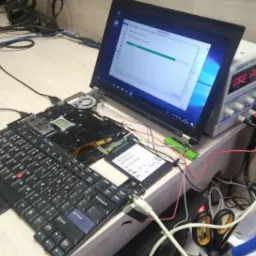 Rozłożony laptop Lenovo ThinkPad z widoczną klawiaturą, płytą główną i podłączonym dyskiem SSD, obok zasilacz laboratoryjny z wyświetlaczem i router Zyxel, sugerujący proces naprawy lub testowania.