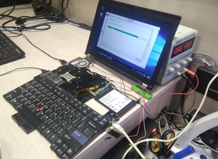 Rozłożony laptop Lenovo ThinkPad z widoczną klawiaturą, płytą główną i podłączonym dyskiem SSD, obok zasilacz laboratoryjny z wyświetlaczem i router Zyxel, sugerujący proces naprawy lub testowania.