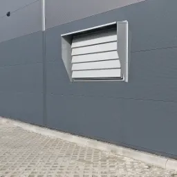 Szary budynek przemysłowy z zamontowaną, białą kratką wentylacyjną z żaluzjami, widoczne także elementy elewacji i chodnik z kostki brukowej.