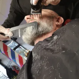 Przycinanie brody siwemu mężczyźnie elektryczną maszynką w salonie, widoczne włosy na pelerynie i charakterystyczny słupek fryzjerski w tle.