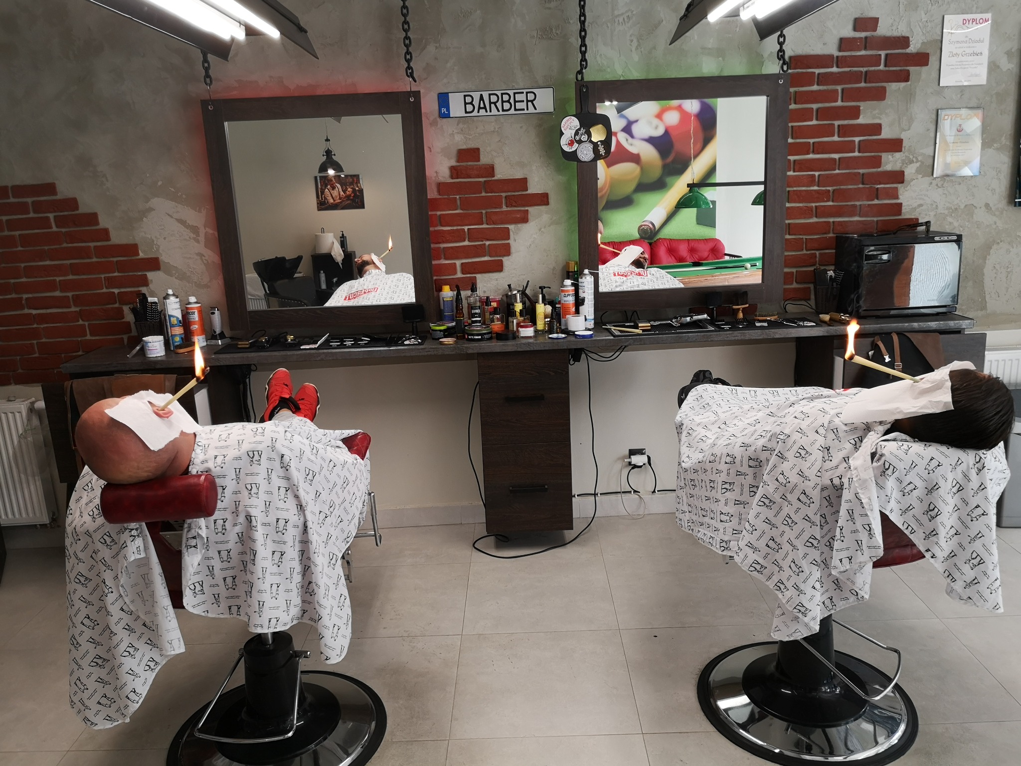 Salon fryzjerski w Koszalinie: Dwa stanowiska z klientami przygotowanymi do golenia brzytwą w ogniu, widok na lustra z odblaskiem wnętrza i dekoracyjną ścianą z cegły.