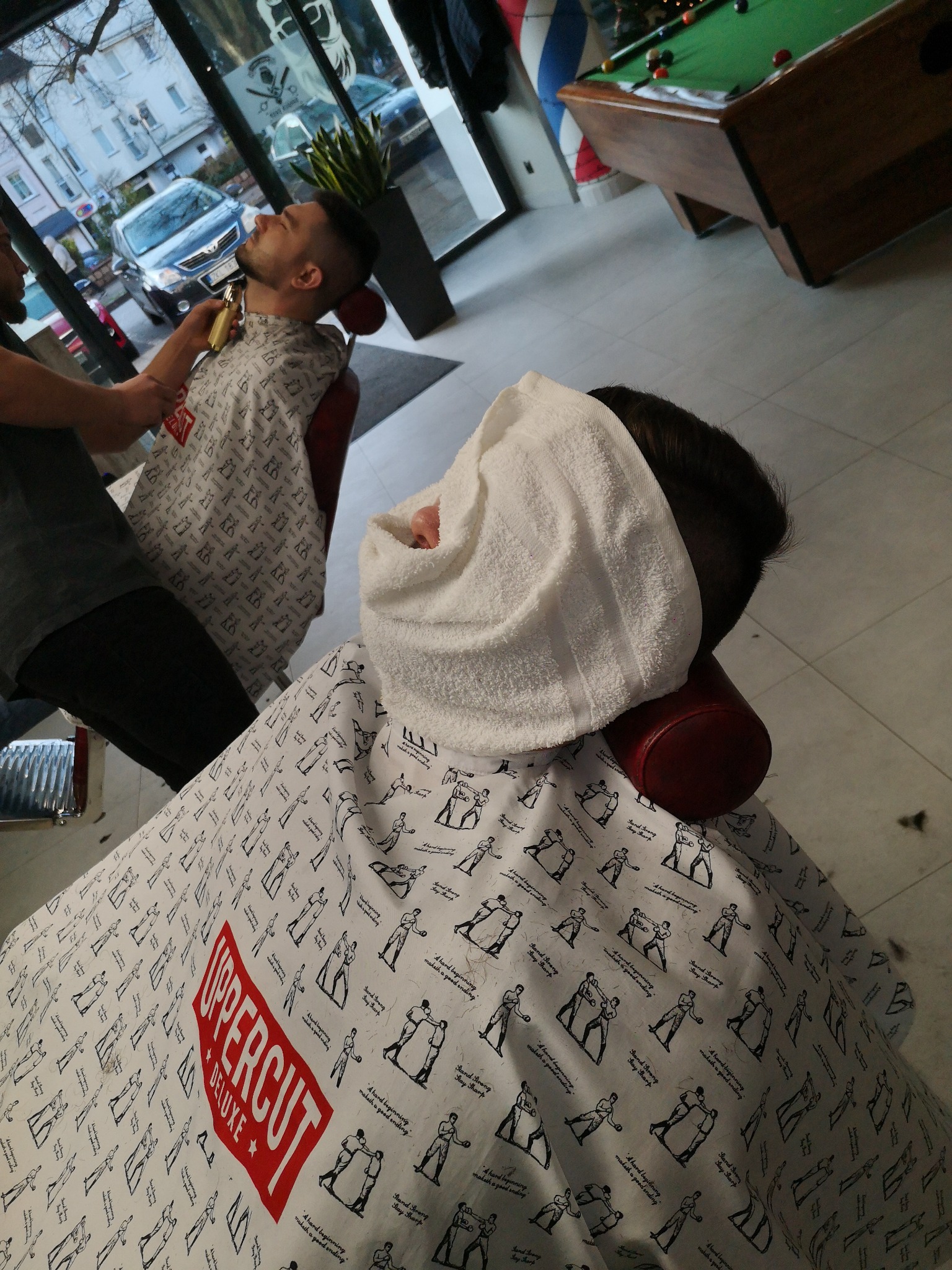 Mężczyzna z zakrytą twarzą ręcznikiem siedzi w fotelu fryzjerskim, na nim peleryna z nadrukiem sylwetek bokserów, w tle fryzjer używa maszynki do strzyżenia włosów innemu klientowi, widoczny stół...