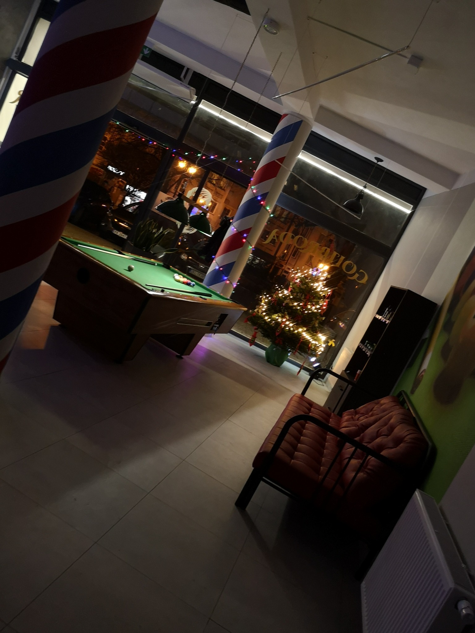 Wnętrze lokalu z bilardem, kolumną barber shop i choinką świąteczną przy oknie.