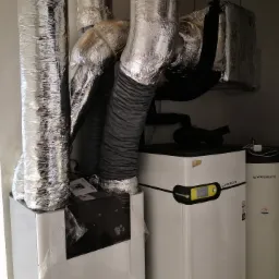 Dwa białe urządzenia AERIS Next i WATERKOTTE, połączone srebrnymi i czarnymi rurami wentylacyjnymi w pomieszczeniu technicznym.