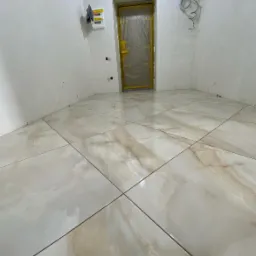 Wykończona podłoga z płytek imitujących marmur w jasnym pomieszczeniu, zabezpieczone drzwi folią ochronną, widoczna skrzynka elektryczna i wystające przewody.