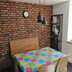 Ceglana ściana w kuchni z drewnianym stołem przykrytym kolorowym obrusem w kratę, nad stołem wiszą dwie lampy w stylu industrialnym, a na ścianie wisi zegar.