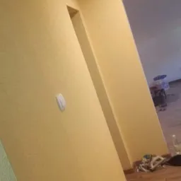 Żółta ściana po nałożeniu gładzi, widoczny włącznik światła i fragment podłogi z paneli, w tle widoczne inne pomieszczenie z elementami wyposażenia.