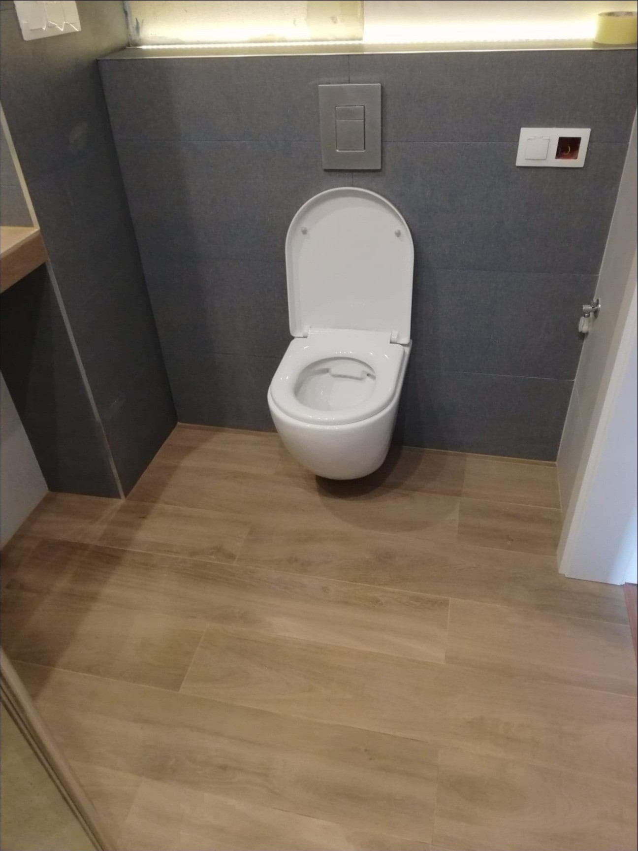 Wisząca toaleta z otwartą klapą na tle szarych płytek ściennych i podłogi imitującej drewno, widoczny przycisk spłuczki i gniazdko elektryczne.