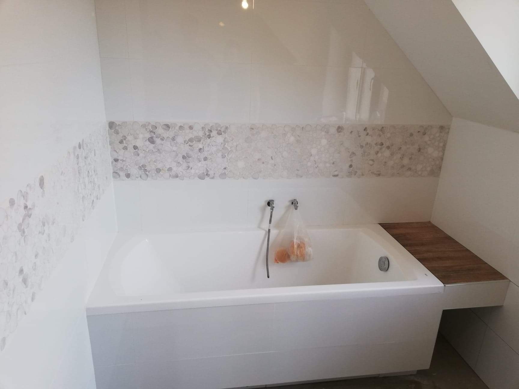 Nowoczesna biała wanna w łazience na poddaszu, z obudową wykończoną płytkami imitującymi kamień rzeczny i drewnianym blatem po prawej stronie; widoczne baterie i worek foliowy z pomarańczowymi...
