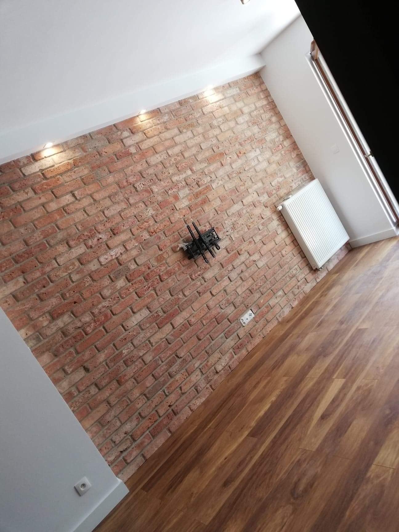 Ceglana ściana z zamontowanym uchwytem na telewizor, biały grzejnik i drewnopodobna podłoga w jasnym, nowoczesnym wnętrzu.