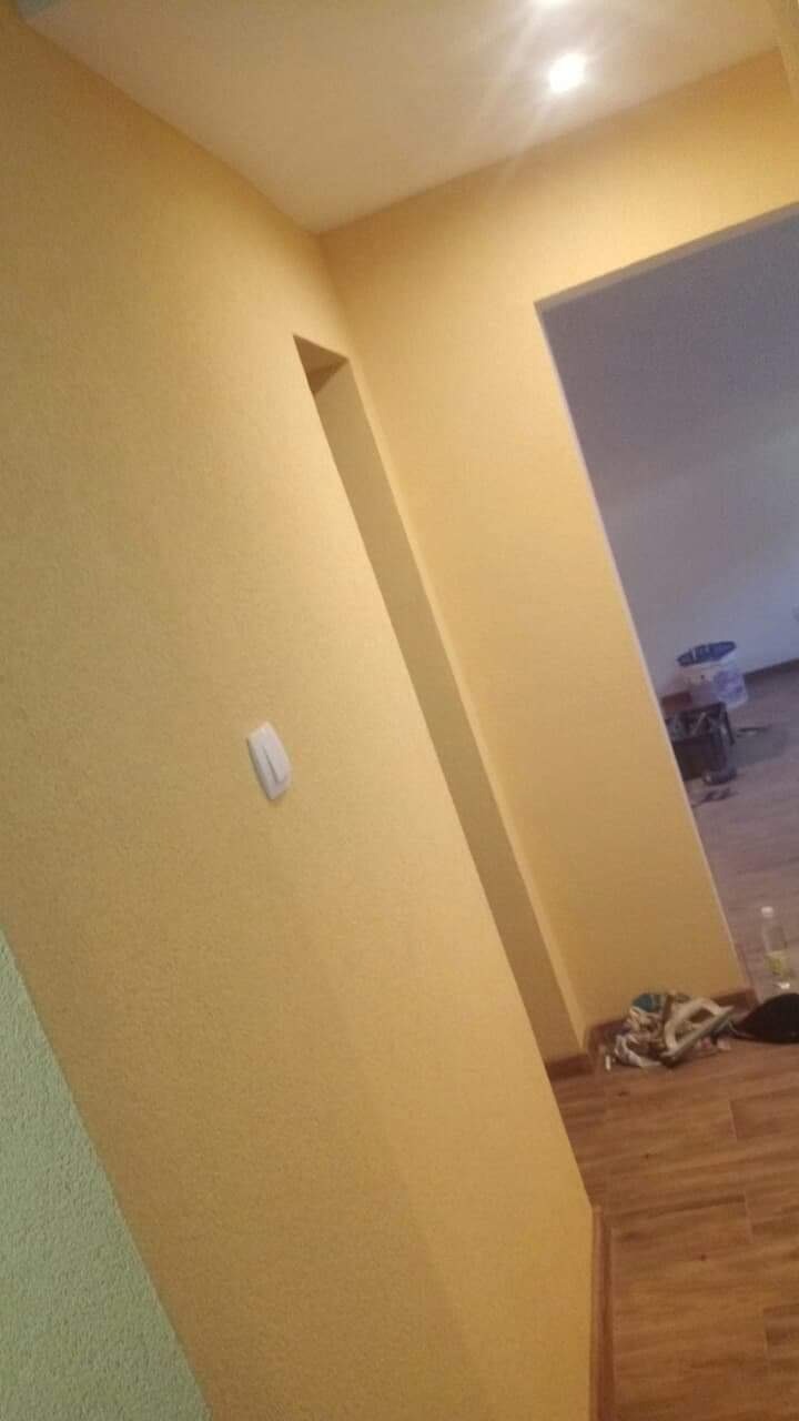 Żółta ściana po nałożeniu gładzi, widoczny włącznik światła i fragment podłogi z paneli, w tle widoczne inne pomieszczenie z elementami wyposażenia.