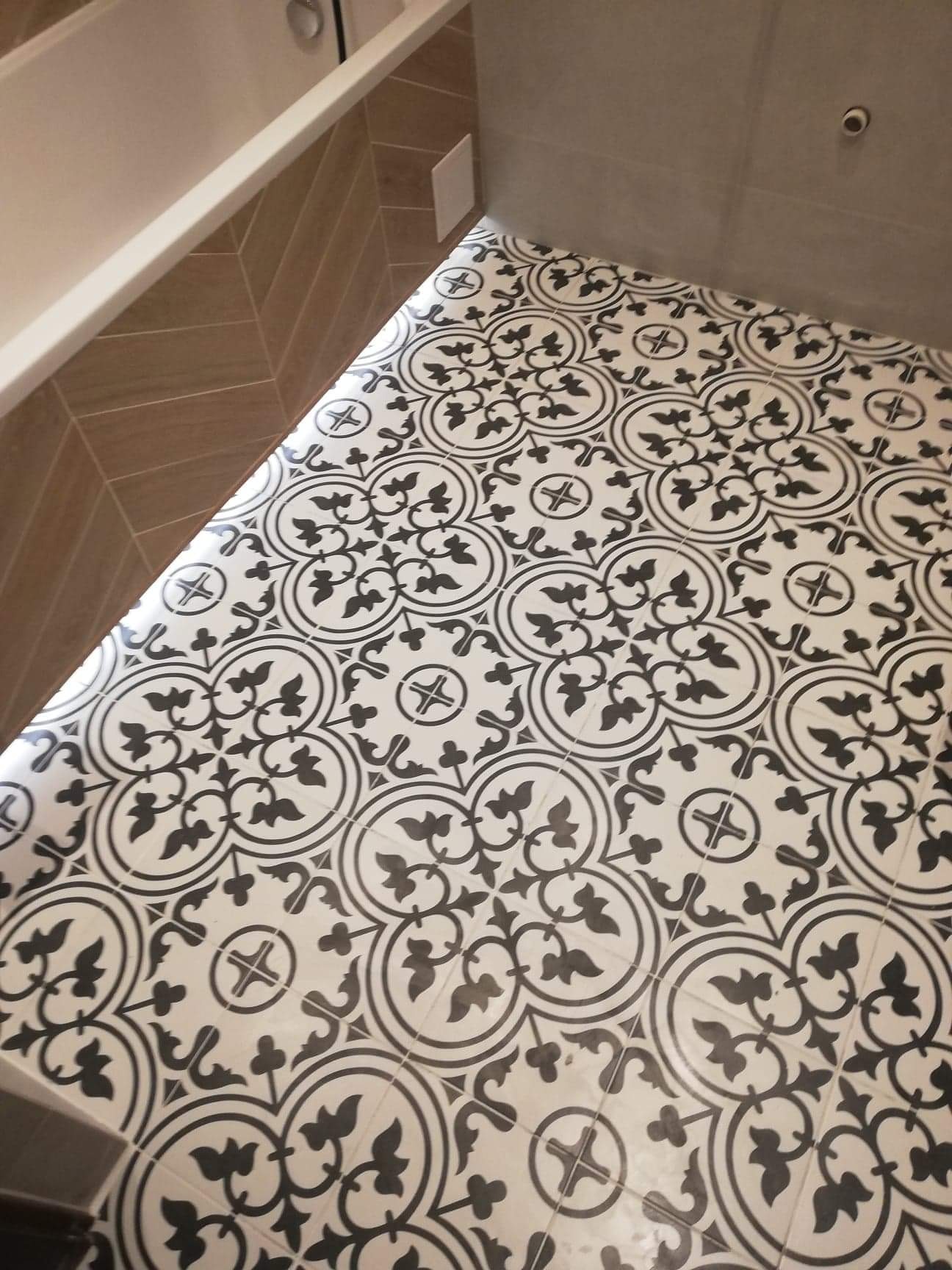 Ułożona podłoga w łazience z płytek w stylu retro, biało-czarne wzory z motywami roślinnymi i geometrycznymi, widoczny fragment wanny i ściany wyłożonej brązowymi płytkami w jodełkę.