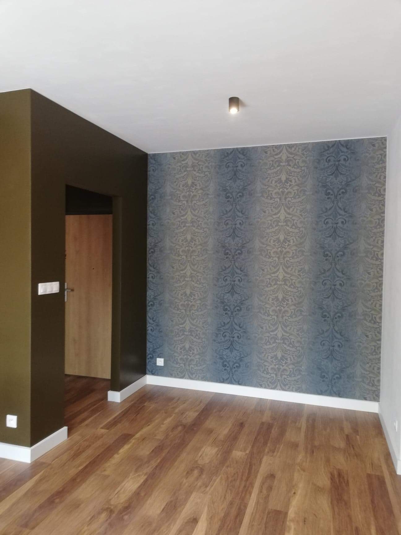 Pomalowane na oliwkowo ściany, panele podłogowe imitujące drewno i ściana z tapetą w ornamenty w wyremontowanym pokoju z zamontowaną lampą sufitową i listwami przypodłogowymi.