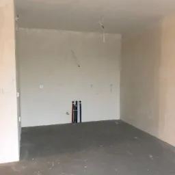 Surowe wnętrze pokoju w trakcie prac wykończeniowych, ściany z widocznymi śladami tynku, wystające rury instalacyjne przy ścianie, betonowa podłoga i wisząca żarówka.