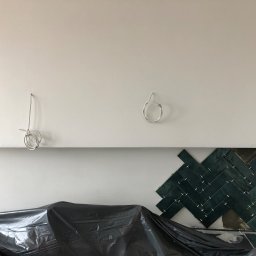 REMMEL Sp. z o.o. - Montaż ciemnozielonych płytek w jodełkę na ścianie, częściowo zakrytych folią ochronną, z widocznymi przewodami elektrycznymi na górze ściany.