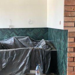 REMMEL Sp. z o.o. - Wnętrze w trakcie wykończenia: ściana z zielonej, ułożonej w jodełkę glazury kontrastuje z fragmentem ściany z cegły, elementy zakryte folią ochronną, widoczne wystające przewody elektryczne.