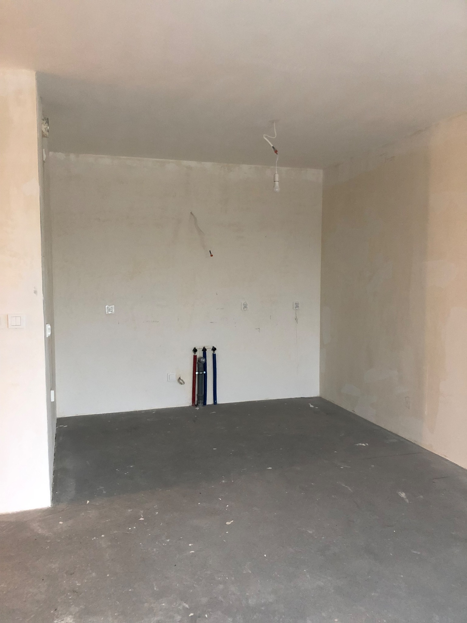 Surowe wnętrze pokoju w trakcie prac wykończeniowych, ściany z widocznymi śladami tynku, wystające rury instalacyjne przy ścianie, betonowa podłoga i wisząca żarówka.
