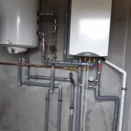 Biały piec gazowy i zasobnik ciepłej wody użytkowej zamontowane na szarej ścianie z widocznymi rurami instalacji centralnego ogrzewania i wodnej, dodatkowo widoczne napisy na ścianie.