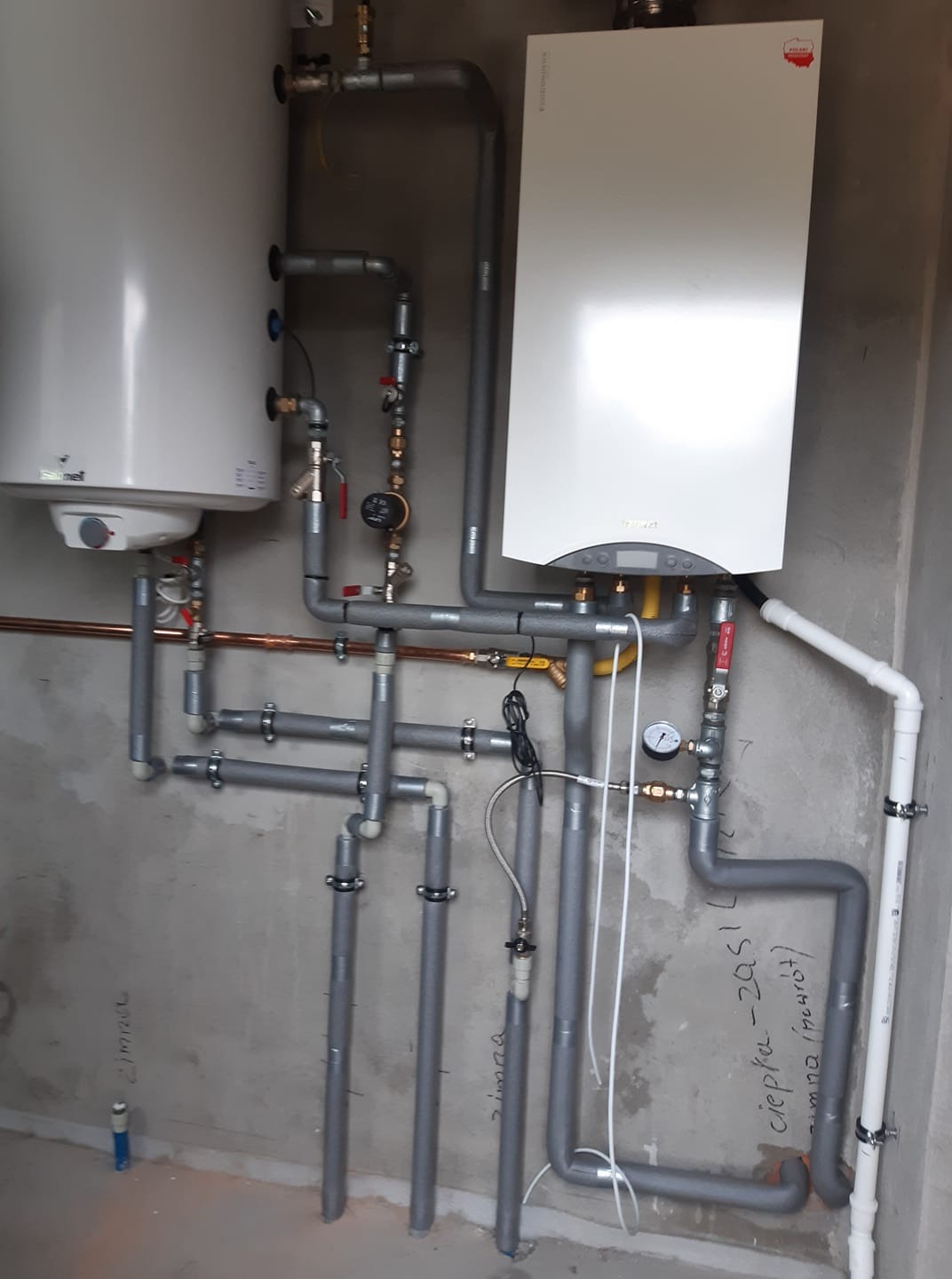 Biały piec gazowy i zasobnik ciepłej wody użytkowej zamontowane na szarej ścianie z widocznymi rurami instalacji centralnego ogrzewania i wodnej, dodatkowo widoczne napisy na ścianie.